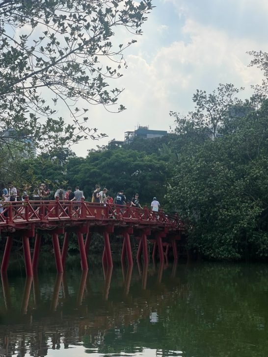 Puente rojo sobre agua con personas y vegetación alrededor.