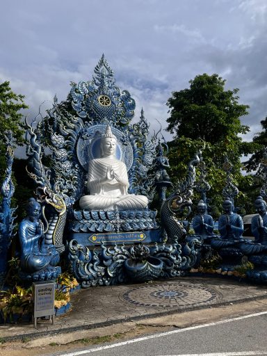 Estatua de Buda blanco rodeada de figuras azules y vegetación. Cielo nublado.