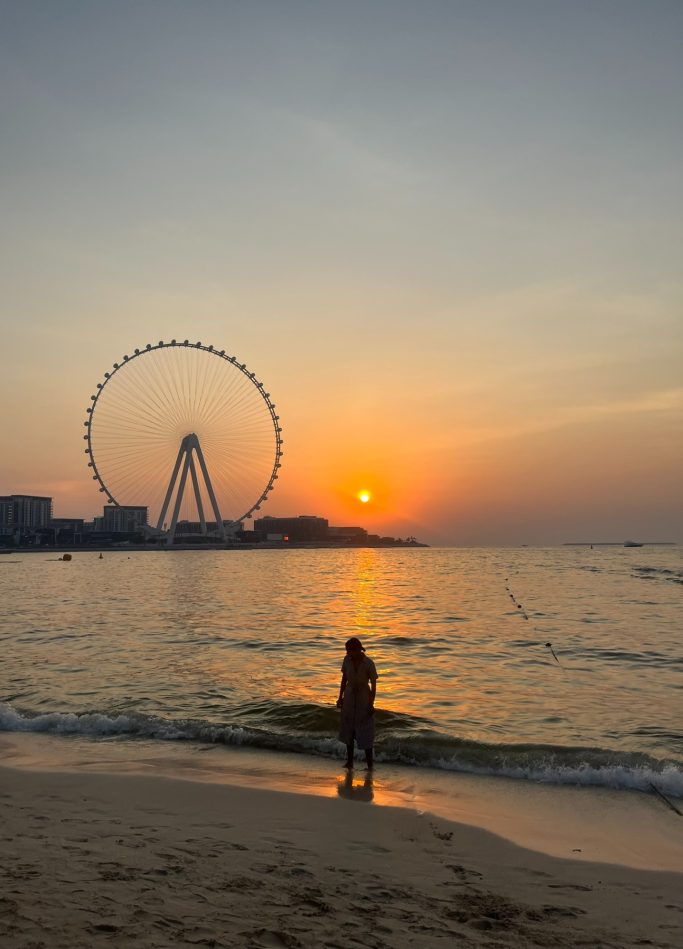 Atardecer en la playa con una persona de pie frente a una noria en el horizonte.