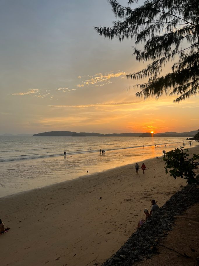 Atardecer en la playa, con personas disfrutando del agua y palmeras al fondo.