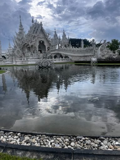 Templo blanco con reflejo en el agua, rodeado de nubes y vegetación.