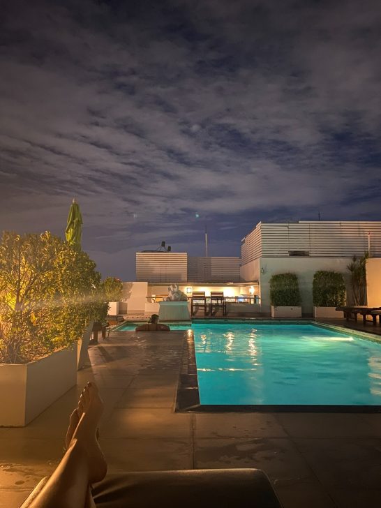 Piscina iluminada por la noche, rodeada de plantas y edificios. Cielo nublado.