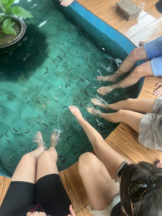 Personas sentadas al borde de una piscina con peces que nadan alrededor de sus pies.