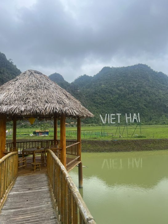 Cabaña de bambú junto a un lago, con montañas al fondo y el texto "VIET HÀ".