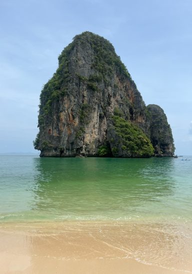 Formación rocosa alta sobre el agua, rodeada de playa y vegetación.