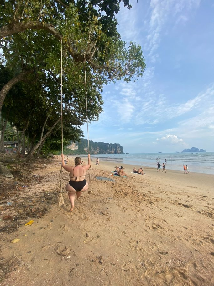 Persona en un columpio en la playa, con mar y montañas al fondo bajo un cielo despejado.