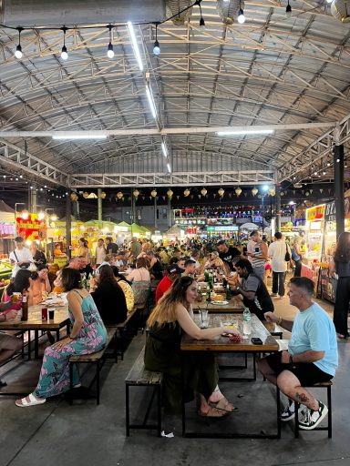 Mercado animado con gente sentada en mesas, disfrutando de comida y ambiente festivo.