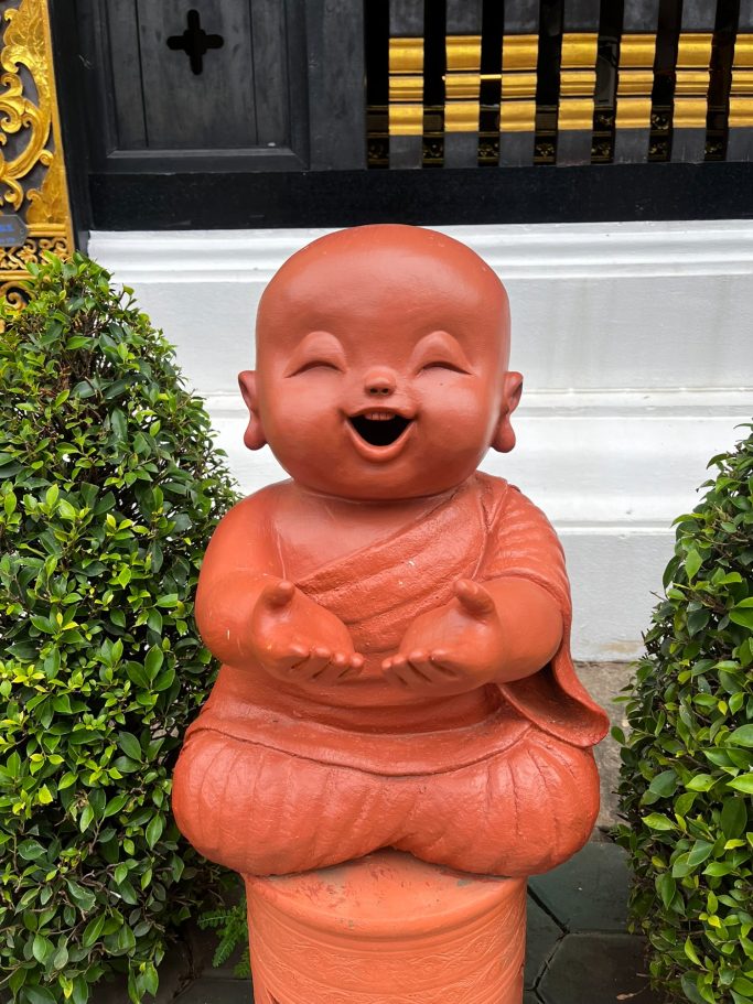 Estatua de un niño sonriente, vestido de monje, rodeado de plantas.