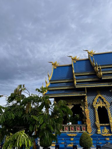Techo azul con detalles dorados, nubes grises en el fondo y vegetación al frente.
