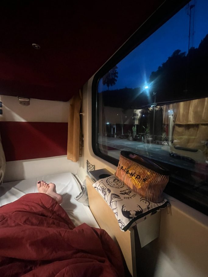 Cama en un vagón de tren con luz suave, ventana que muestra un paisaje nocturno.