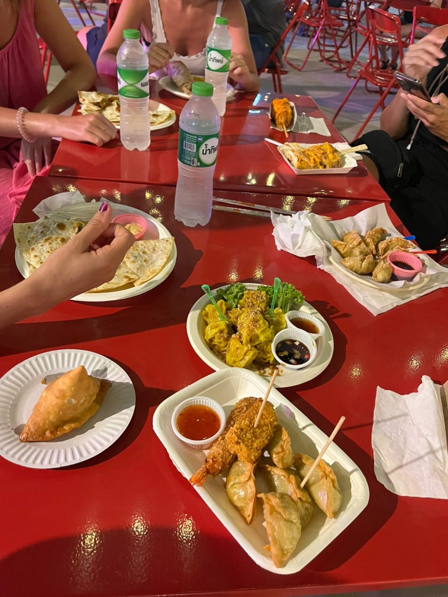 Variada comida en bandejas sobre una mesa roja, con bebidas y gente comiendo alrededor.