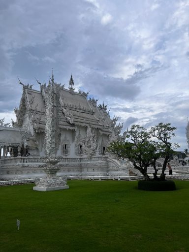 Templo blanco con detalles elaborados y un cielo nublado de fondo. Jardín bien cuidado.