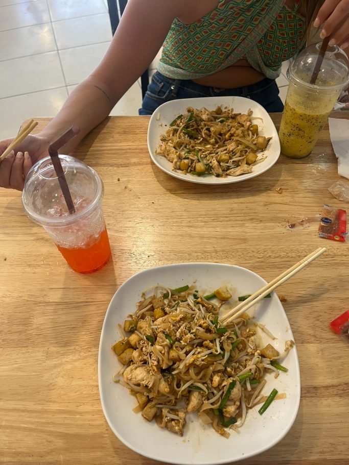 Platos de comida asiática con fideos, verduras y dos bebidas en mesas de madera.