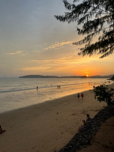 Atardecer en la playa, con personas caminando y olas suaves. Árboles al fondo.