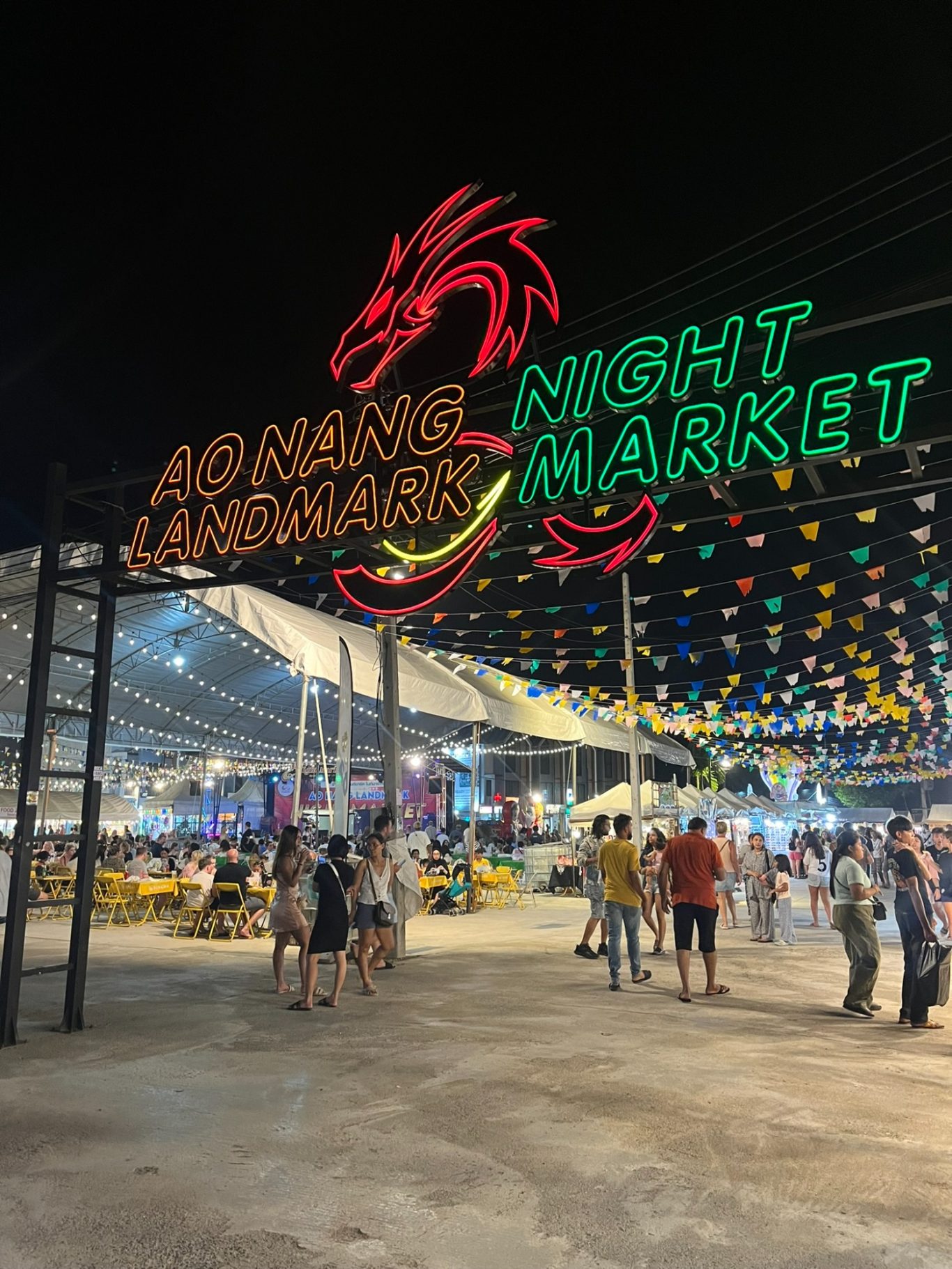 Mercado nocturno con un letrero de dragón rojo y luces coloridas. Gente caminando.